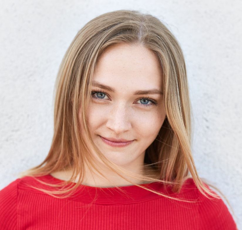 Serene young woman with long blonde hair and blue eyes, smiling gently, wearing a red top against a white background.