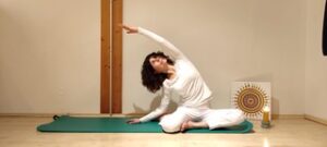 A woman in white clothing performs a seated side-bend yoga pose on a green mat in a softly lit room with a candle.