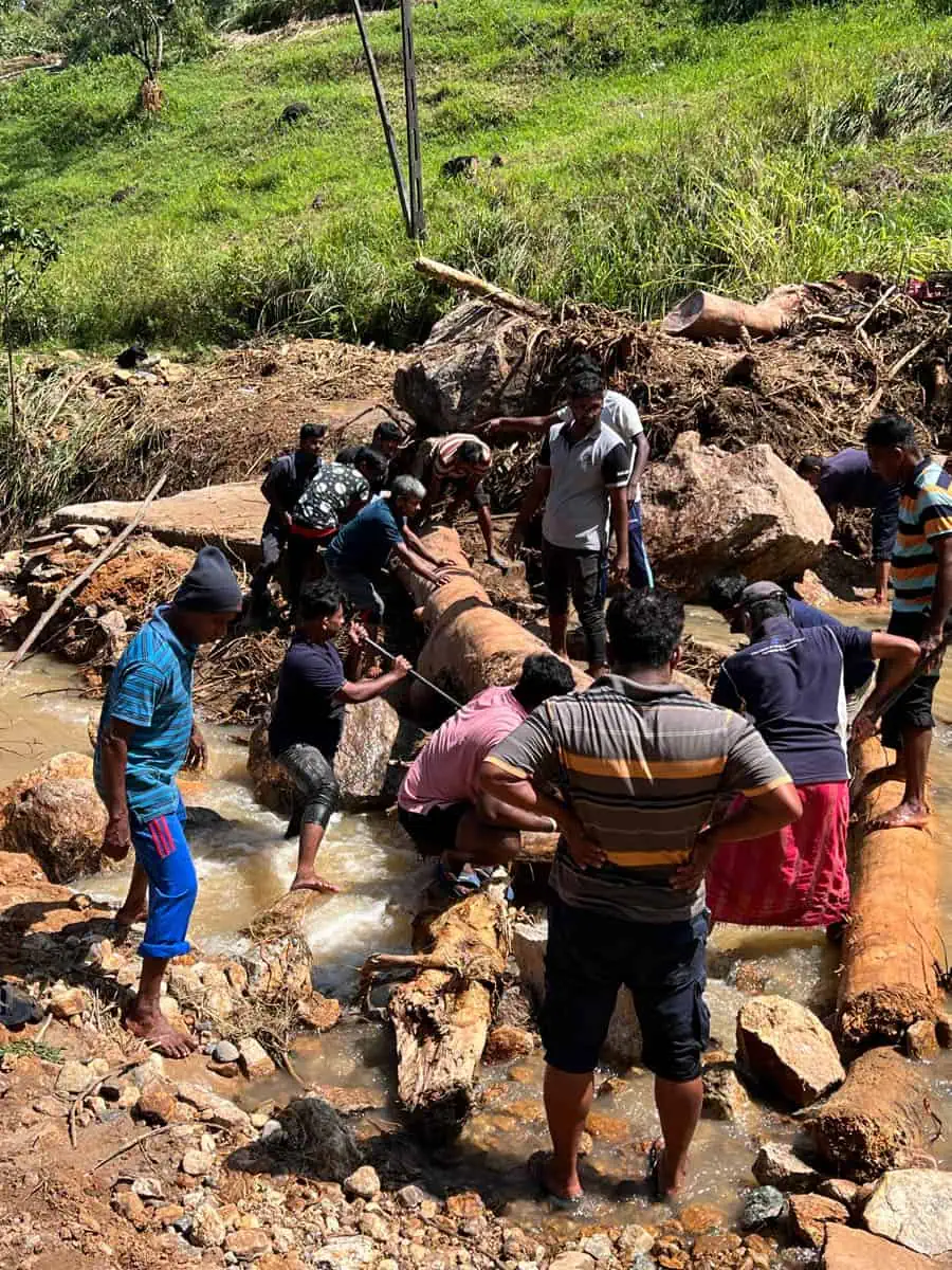 A team of local workers maneuvers a large, wrapped pipe across a rocky stream in a green valley.