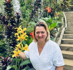A woman smiles while touching a stalk of bright yellow flowers in a lush tropical garden with stone steps.